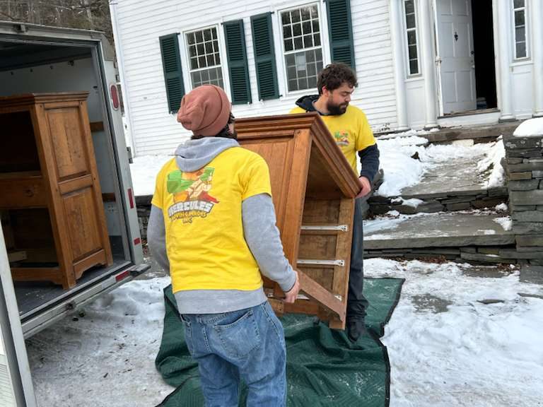 Two Hercules movers carrying a wooden cabinet out of the back of a truck across a snowy driveway