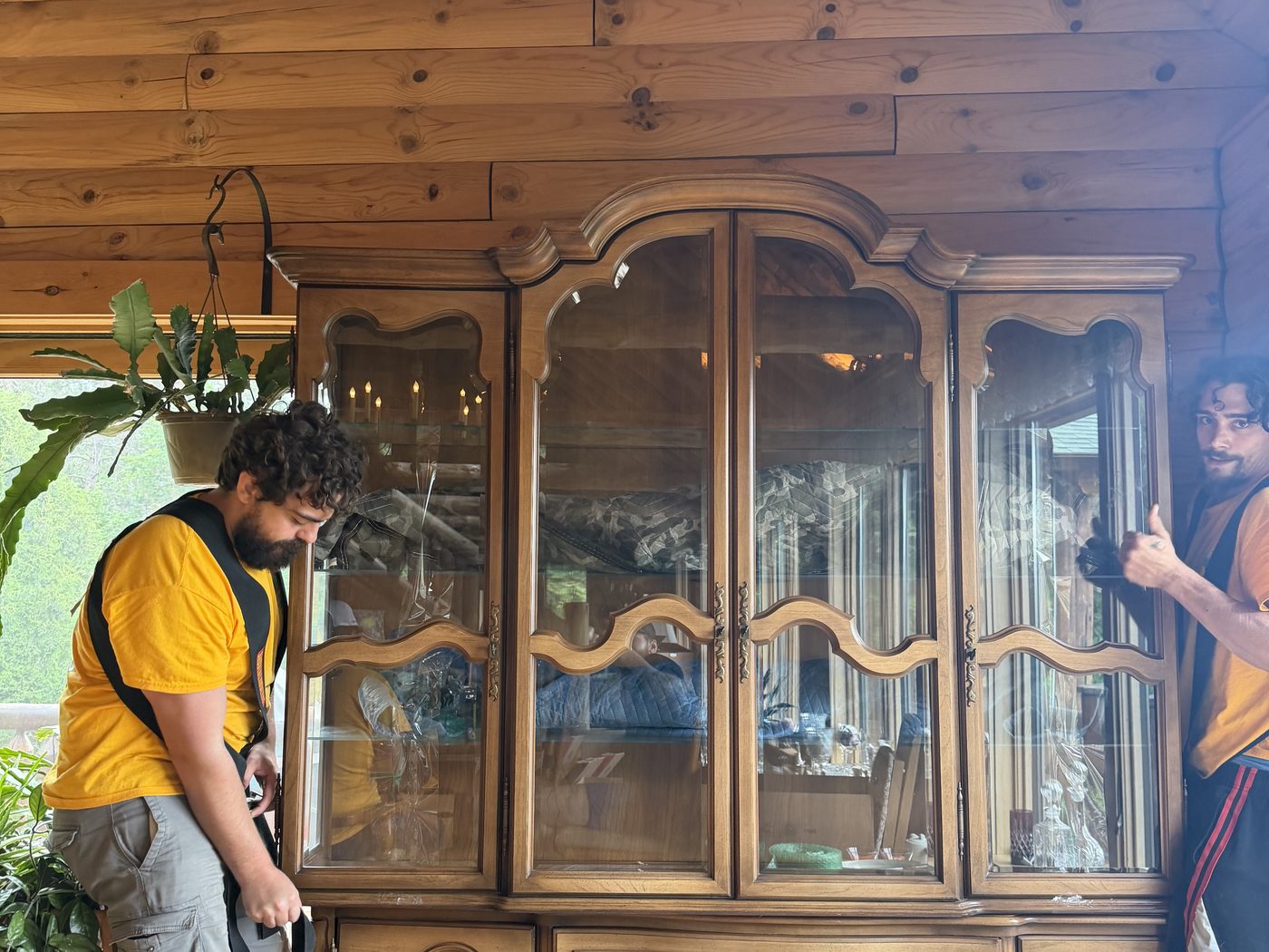 Two Hercules movers lifting a glass-front china cabinet on shoulder straps in a log home
