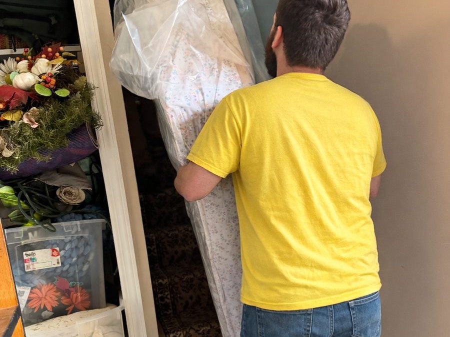 A Hercules mover in a yellow shirt carrying a mattress wrapped in plastic through a narrow doorway toward a staircase