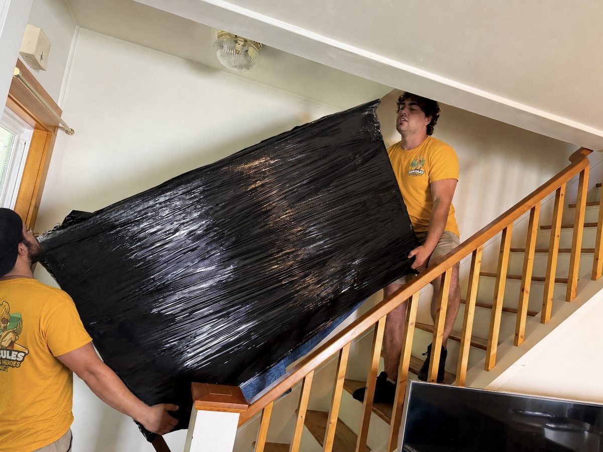 Two Hercules movers carrying a wrapped dresser down an exterior staircase