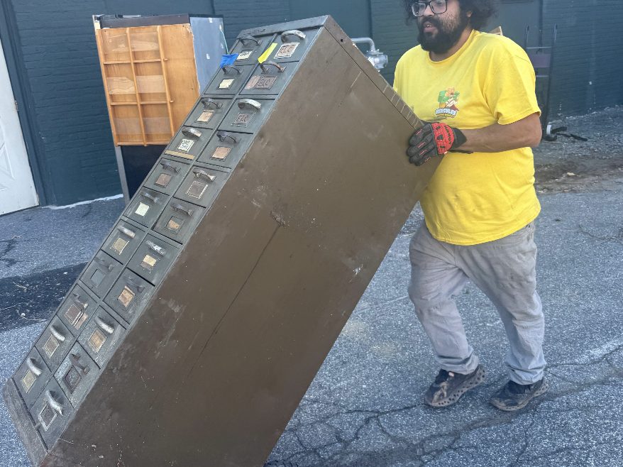 A Hercules mover wheeling a metal file cabinet on a dolly across a parking lot