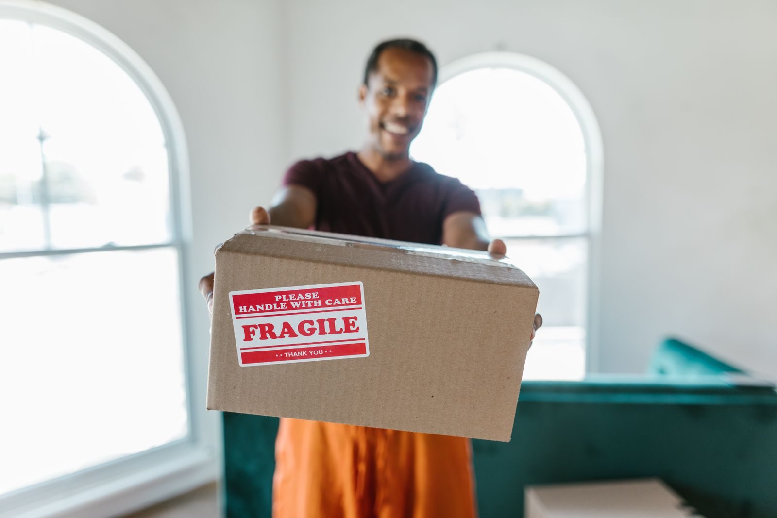 A smiling mover holding out a cardboard box with a red fragile label in a bright room