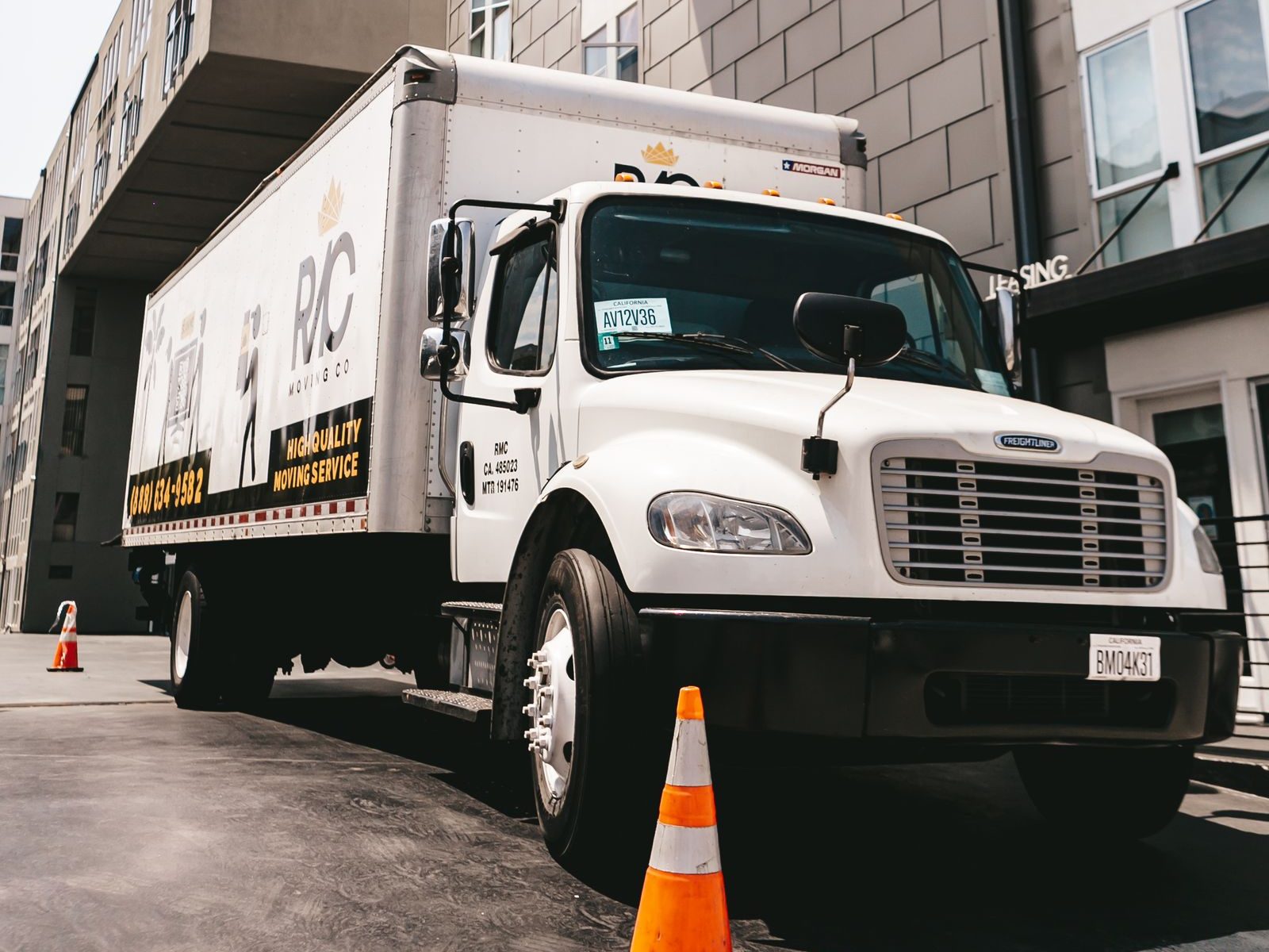 A white moving truck parked outside a modern apartment building with an orange cone beside it