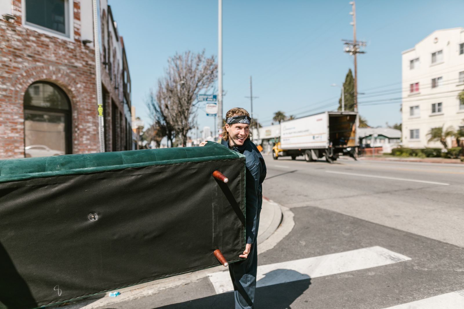A mover in a dark t-shirt carrying a long green sofa across a sunlit city street