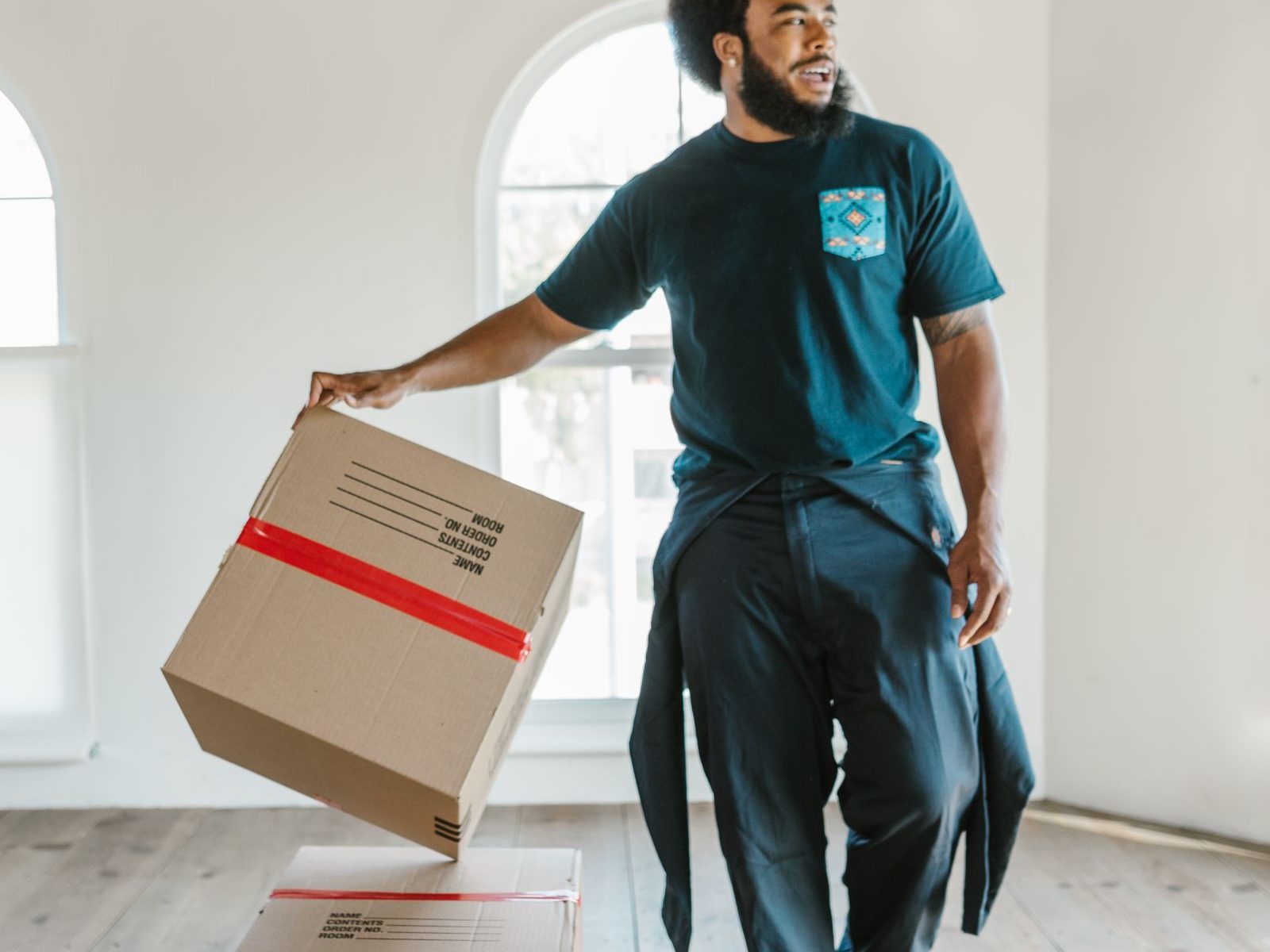 A mover in a dark t-shirt lifting a taped cardboard box in a bright empty room