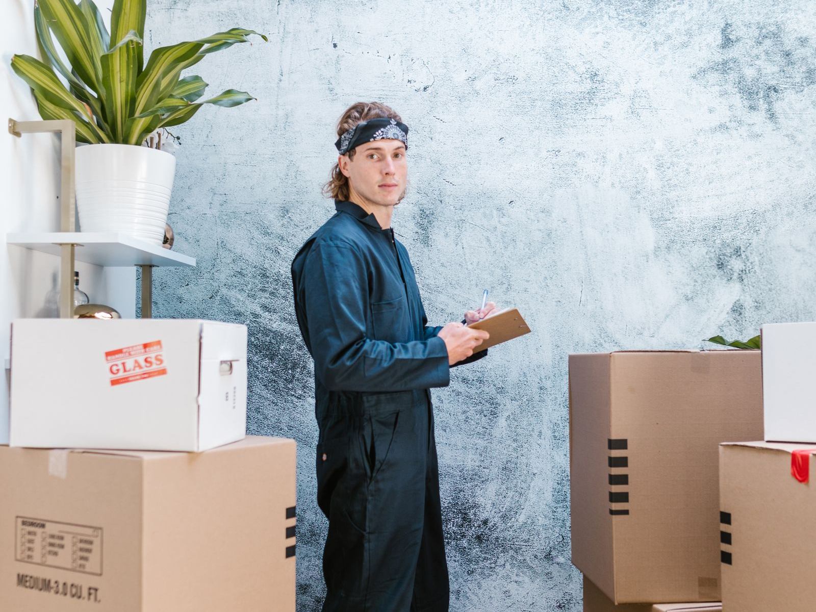 A mover in dark coveralls and a bandana checking a clipboard beside stacked cardboard boxes
