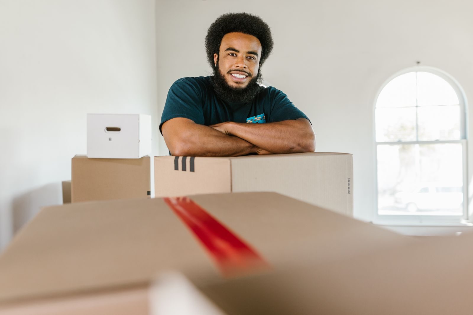 A smiling mover in a dark t-shirt leaning on a stack of taped moving boxes in a bright room