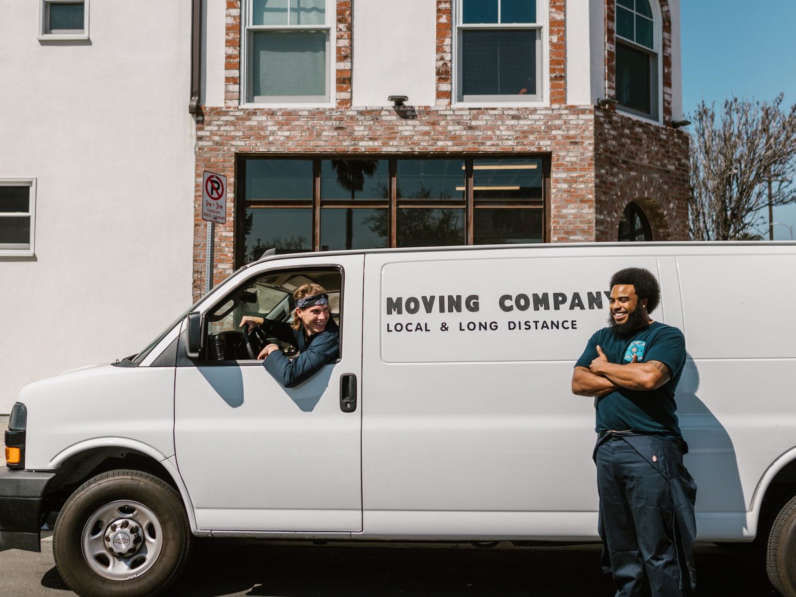 A white cargo van lettered Moving Company parked outside a brick building, one mover at the wheel and another standing beside it