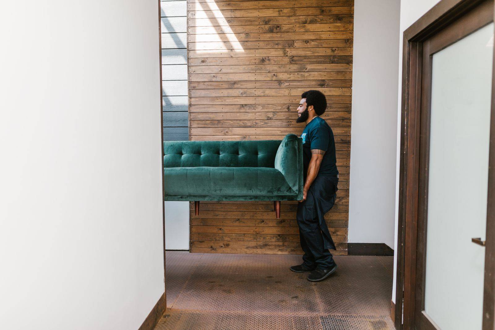 A mover in a dark t-shirt carrying a green velvet sofa through a doorway