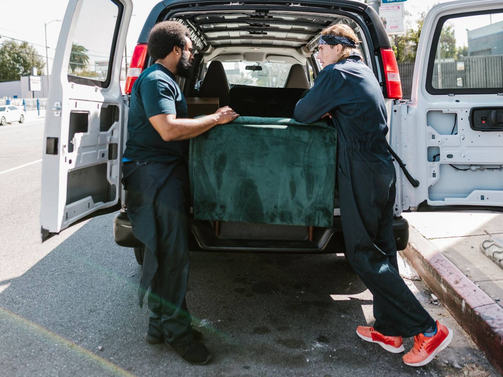 Two movers loading a green sofa into the back of a white cargo van on a city street