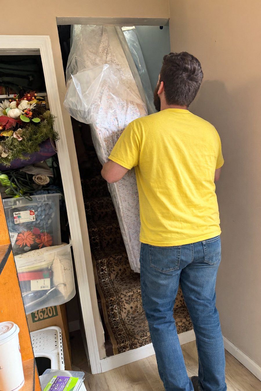 A Hercules mover carrying a mattress wrapped in plastic through a narrow doorway toward a staircase