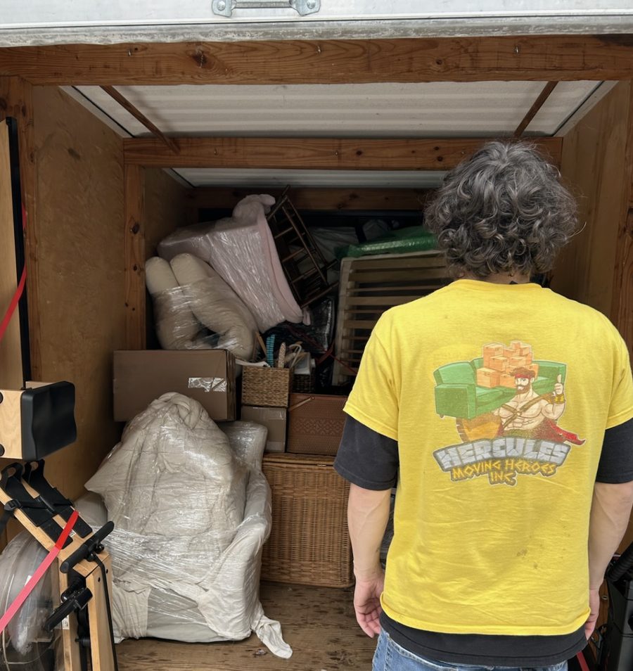 Logan, back to the camera in a Hercules shirt, in front of a truck loaded with wrapped furniture and boxes