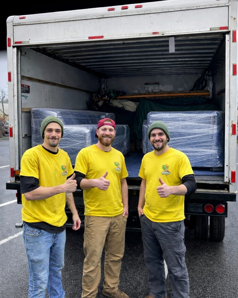 Three Hercules movers in matching yellow shirts giving a thumbs up in front of a truck loaded with wrapped furniture